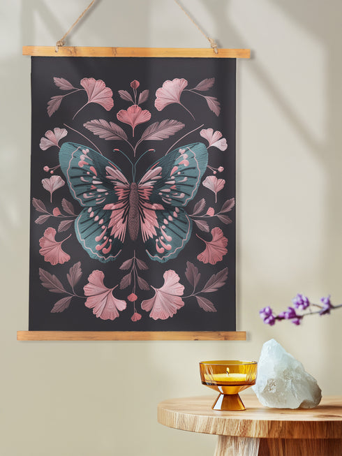Hanging tapestry featuring a teal and pink butterfly surrounded by ginkgo-like leaves on a black background. The textile is suspended from wooden dowels with twine, creating detailed 3D embroidery-like texture.